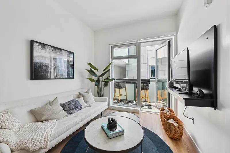 Bright Living Room With White Sofa, Wall-Mounted Television, Round Coffee Table, Wood Flooring, And Private Balcony.